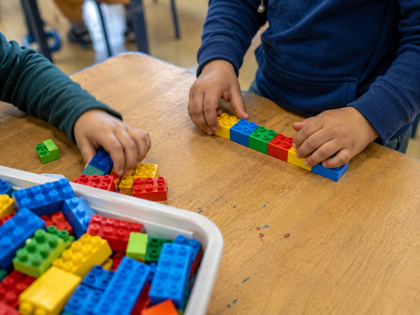 Young children's hands arranging six coloured DUPLO-style bricks into a pattern on a classroom table